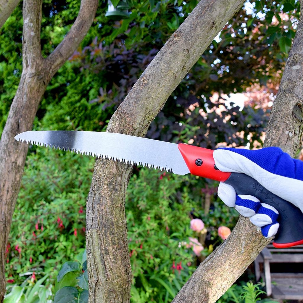 A pruning saw, with a red and black handle being operated by a person with blue and white gloves, cutting into a thick branch